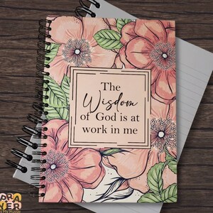 以下が含まれることがあります： ピンクとグリーンの花柄のデザインのノート。"The Wisdom of God is at work in me"というテキストが書かれています。