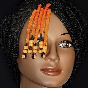 May include: A mannequin head with a black braided wig and orange and yellow beaded accents. The mannequin is wearing a black turtleneck sweater against a black background. The wig features a side-swept style with the beaded accents.