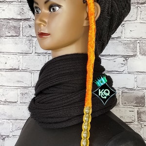 May include: A black beanie with a long orange and yellow beaded hair extension. The beanie is made of a soft, knitted fabric and has a slouchy fit. The hair extension is attached to the beanie with a small clip.