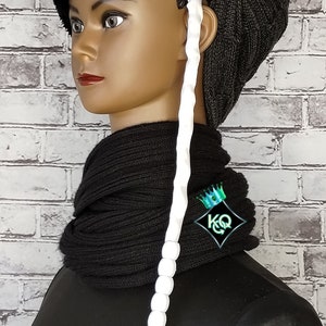 May include: A black knit beanie with a white beaded braid attached. The beanie is worn on a mannequin with a black knit scarf.
