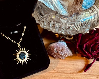 May include: A gold-toned necklace featuring a sunburst pendant with a dark circular center and a halo of small clear stones. The pendant hangs from a delicate gold chain, presented inside a black velvet-lined jewelry box.