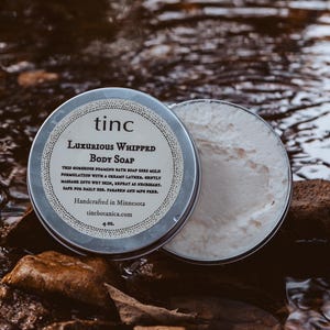 May include: A silver tin with a white label that reads "tinc LUXURIOUS WHIPPED BODY SOAP" and contains a white whipped body soap. The tin is sitting on a bed of rocks and water.
