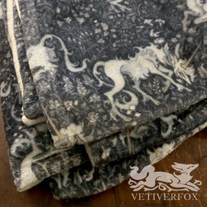 Könnte beinhalten: Eine gefaltete, dunkelgraue Decke mit einem weißen Einhorn-Design. Die Decke weist ein sich wiederholendes Muster aus Einhörnern und floralen Elementen auf. Die Ränder sind mit einem weißen, flauschigen Rand versehen. Das Vetiverfox-Logo ist unten rechts sichtbar.