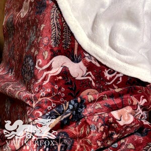 May include: A plush, red blanket with a detailed tapestry design featuring a white greyhound, floral patterns, and foliage. The blanket has a soft, white, fluffy border. The brand name "VEITVERFOX" is visible in the bottom left corner.