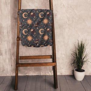 Puede incluir: Una manta azul oscuro con un diseño celestial de soles dorados, lunas crecientes, estrellas y nubes burdeos. La manta está colocada sobre una escalera de madera. Una planta en maceta está a la derecha.