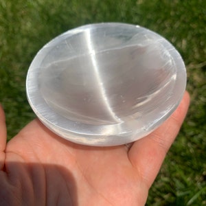 May include: A clear, round selenite bowl with a smooth, polished surface. The bowl is held in a hand, with the green grass in the background. The bowl is approximately 4 inches in diameter.