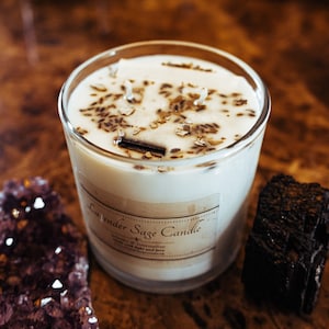 May include: A clear glass candle with a white wax interior, labeled "Lavender Sage Candle." The candle is topped with dried lavender and wood pieces. A purple crystal and a black stone are on either side of the candle.