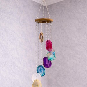 Op de afbeelding: Een hangende windgong met een houten ring en zilveren kettingen. De kettingen hangen kleurrijke, gepolijste agaat schijfjes in tinten blauw, paars, roze en geel. De agaat schijfjes variëren in grootte en vorm, wat een decoratieve uitstraling creëert.