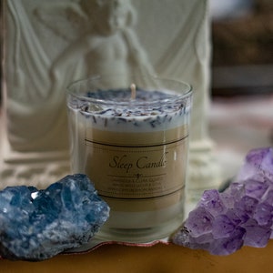 May include: A clear glass candle with a label that reads "Sleep Candle". The candle is filled with a white wax base, topped with lavender and dried herbs. Two crystals, one blue and one purple, flank the candle.