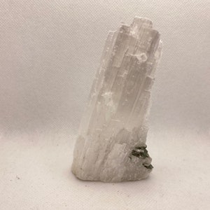 May include: A translucent selenite crystal, pale off-white in colour, with a layered, fibrous texture. A small cluster of dark green minerals is at the base of the crystal. The selenite is a mineral specimen.