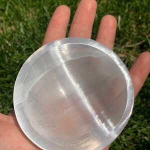 May include: A clear, round selenite charging plate. The plate is held in a hand, with the green grass in the background. The plate has a smooth, reflective surface and is used for cleansing and charging crystals.