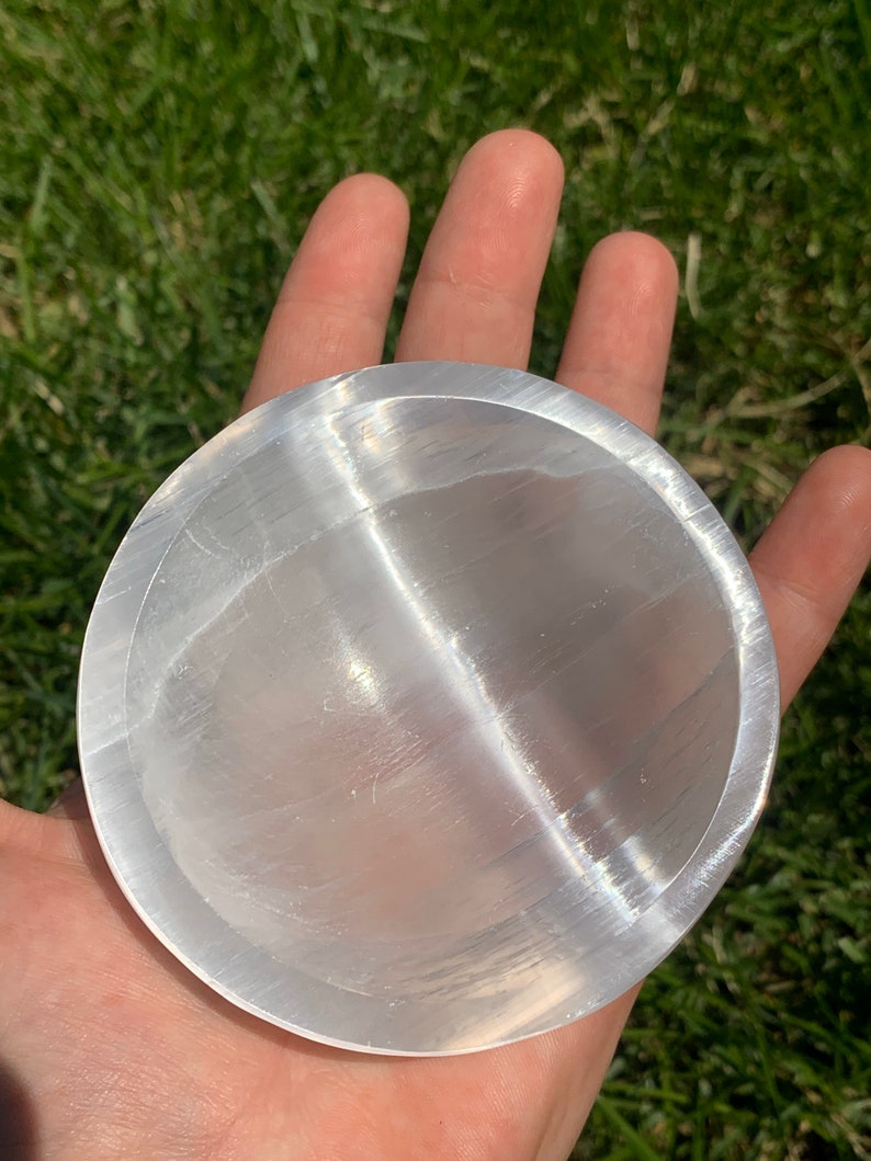May include: A clear, round selenite charging plate. The plate is held in a hand, with the green grass in the background. The plate has a smooth, reflective surface and is used for cleansing and charging crystals.