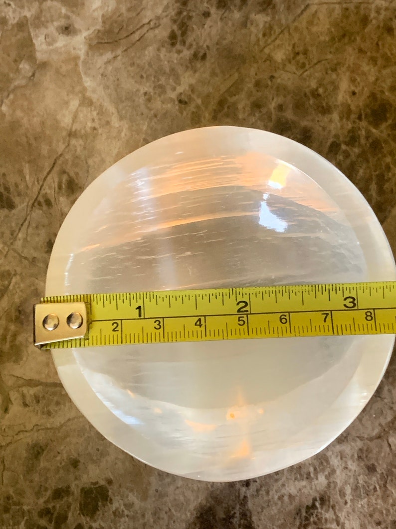 May include: A round, white selenite bowl with a translucent appearance. A yellow measuring tape is placed across the bowl, showing a diameter of approximately 7 inches.
