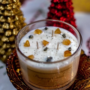 May include: A clear glass candle holder with a white candle, adorned with yellow and gray stones. The candle has three wicks and a label that reads "Prosperity". The candle sits on a decorative, ornate, brown plate. Two Christmas trees are in the background.