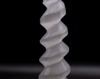 May include: A spiraling, light gray selenite crystal sculpture. The carved stone has a twisted, vertical design with a pointed top and a wide base. The sculpture is set against a black background.
