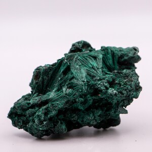 May include: A natural malachite specimen, displaying a deep green hue and a fibrous structure. The stone's surface has a rough, textured appearance, with visible banding. The malachite is presented against a plain white background, highlighting its natural form.