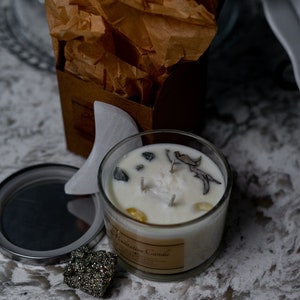 May include: A clear glass Meditation Candle with a white wax interior, adorned with small stones and dried botanicals. The candle has three wicks and a label. A silver lid and a piece of pyrite are next to the candle. A brown box with brown paper is in the background.