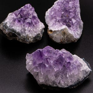 May include: Three amethyst crystal clusters, displaying a rich purple colour and a crystalline structure. The crystals are set against a black backdrop, highlighting their natural texture. The raw, uncut stones vary in size and shape.
