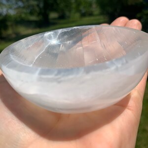 May include: A clear, oval-shaped selenite bowl. The bowl is held in a hand, with the natural light reflecting off its smooth, translucent surface. The bowl's interior is visible, showcasing its carved design. The background is blurred, with green foliage.