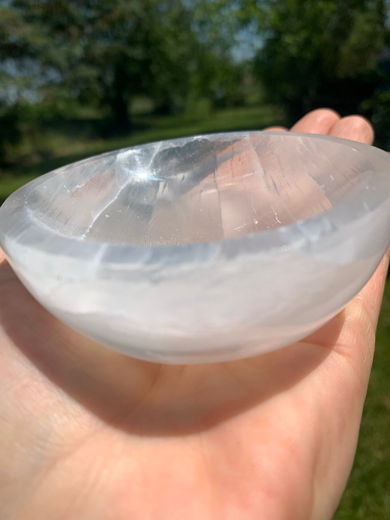 May include: A clear, oval-shaped selenite bowl. The bowl is held in a hand, with the natural light reflecting off its smooth, translucent surface. The bowl's interior is visible, showcasing its carved design. The background is blurred, with green foliage.