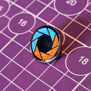 Peut inclure: Une épingle noire et argentée avec un symbole d'ouverture bleu et orange. L'épingle a une finition scintillante.