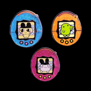 May include: Three colourful enamel pins shaped like a Tamagotchi, a popular virtual pet toy. The pins feature different characters on the screen, including a yellow bunny with black ears, a green frog, and a pink bunny with black ears. The pins are a fun and nostalgic way to show your love for the classic toy.