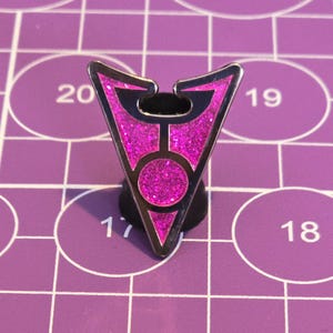 Puede incluir: Un pin de color plateado con un diseño de purpurina rosa. El pin tiene forma de triángulo con un círculo en el centro. El pin está sobre una cuadrícula morada.