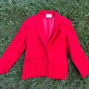 May include: A vibrant red blazer featuring a notched lapel, long sleeves, and a two-button front closure. The garment includes two horizontal flap pockets at the waist and a matching red interior lining with a visible white brand label at the neck.
