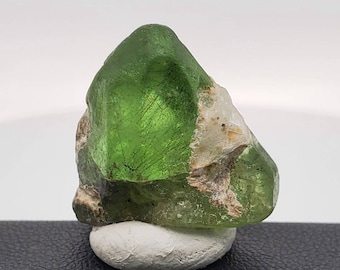 Puede incluir: Un cristal verde con una textura rugosa y natural. El cristal tiene una punta puntiaguda y una base plana. Está parcialmente cubierto de depósitos minerales blancos y marrones.