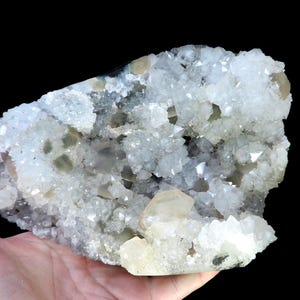 Pode incluir: Um aglomerado de cristais de quartzo em tons de branco, cinza e amarelo pálido. Os cristais têm uma superfície áspera e texturizada e são segurados em uma mão contra um fundo preto. A forma geral é irregular e orgânica.