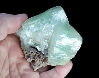 Op de afbeelding: Een hand houdt een grote, doorschijnende, lichtgroene kristalformatie vast. Het kristal heeft een gelaagde structuur met scherpe randen en een ruwe, bruine matrixbasis. De algehele uitstraling is kristallijn en natuurlijk.