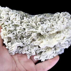 Pode incluir: Close-up de um espécime mineral com formações cristalinas brancas. Os cristais têm uma aparência borbulhante e texturizada, com algumas áreas mais opacas. O fundo é preto.