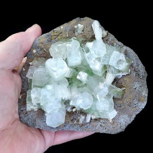 May include: A cluster of pale green crystal formations on a rough, gray-brown rock base. The crystals are translucent and have a geometric shape. The rock has a textured surface. The overall appearance is natural and geological.