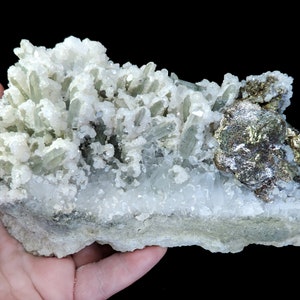Pode incluir: Um aglomerado de cristais de quartzo transparentes com um tom verde, alguns dos quais são alongados e pontiagudos, estão dispostos sobre um leito de cristais de quartzo brancos. Uma grande peça de pirita, um mineral metálico de cor prateada-dourada, está aninhada entre os cristais de quartzo.