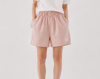 May include: A white short-sleeve t-shirt with a chest pocket paired with dusty pink elastic-waist shorts featuring a small rectangular brand label on the lower left leg and brown strappy sandals. The outfit is completed with a light-colored shoulder bag.