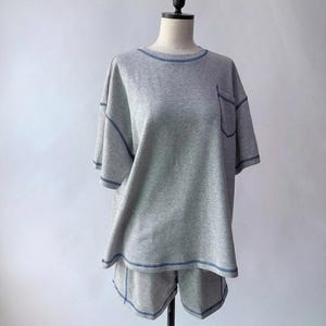 May include: A light grey short-sleeve t-shirt and matching shorts set featuring prominent dark blue contrast stitching along the neckline, sleeve hems, pocket edge, and bottom hem. The t-shirt includes a single chest pocket on the left side.
