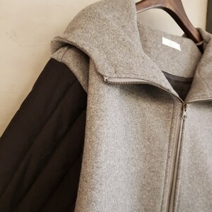 Puede incluir: Una chaqueta gris con capucha y una manga acolchada negra. La chaqueta tiene una cremallera.