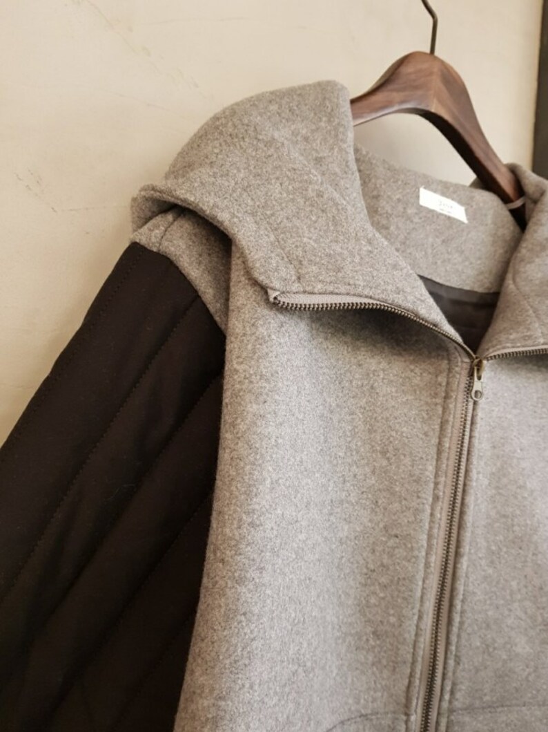 Puede incluir: Una chaqueta gris con capucha y una manga acolchada negra. La chaqueta tiene una cremallera.