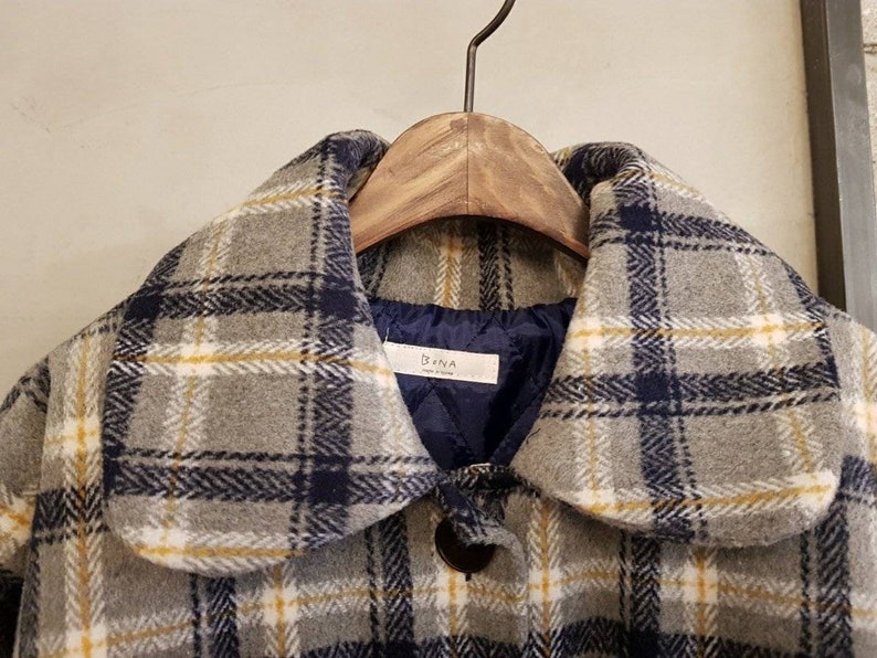 May include: A gray coat featuring a plaid pattern in navy blue, white, and yellow lines. The coat has a rounded collar and a dark blue quilted interior lining. A white label with the text Bona is sewn into the inner neckline of the garment.