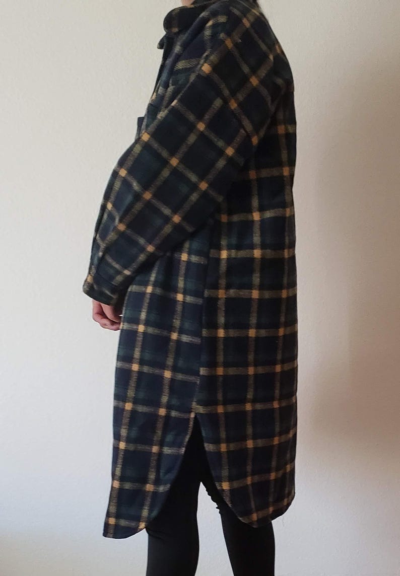 May include: A long-sleeved, knee-length shirt jacket featuring a dark navy blue, forest green, and mustard yellow plaid pattern. The garment has a classic collar, a button-down front, long sleeves with cuffs, and a curved hem with side slits.