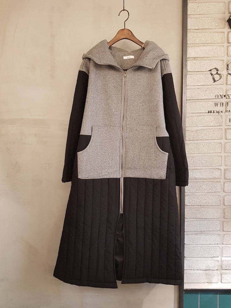 Puede incluir: Una chaqueta larga con capucha gris y negra con cierre de cremallera. La chaqueta tiene un fondo acolchado y dos bolsillos en la parte delantera.