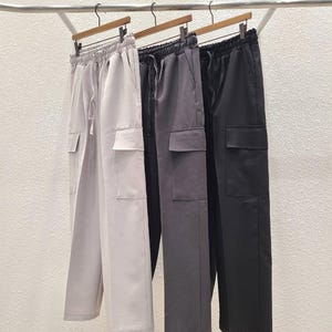 Op de afbeelding: Drie paar cargo broeken hangen aan een rek. De broeken zijn grijs, zwart en donkergrijs.