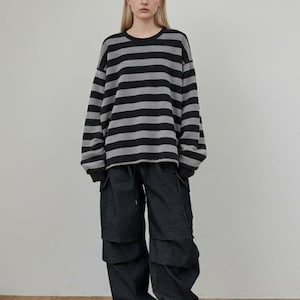 May include: A person wearing a black and gray striped long-sleeve shirt and dark denim cargo pants. The person is wearing red sneakers with white soles. The outfit is casual and modern.