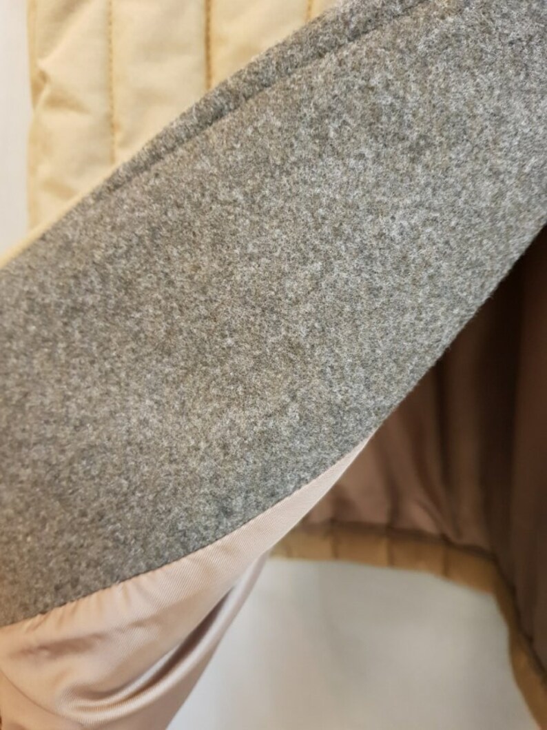 Puede incluir: Primer plano del interior de una chaqueta acolchada beige. El forro es de tela gris claro con un forro liso y sedoso debajo.