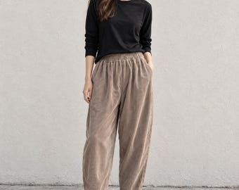 May include: A long-sleeved black crew neck shirt paired with high-waisted, wide-leg corduroy trousers in a light brown taupe color. The trousers feature an elasticized waistband and a textured vertical ribbed pattern, finished with black slip-on shoes.