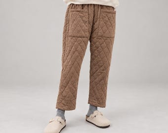 May include: Brown quilted trousers featuring a diamond-stitched pattern, an elasticized waistband, and two large front patch pockets. The pants are paired with grey ribbed socks and light beige slip-on clogs with a single buckle strap detail.