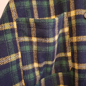 May include: A flannel shirt featuring a dark navy blue, forest green, and bright yellow plaid pattern. The garment includes a single patch pocket on the front and a dark button visible near the top edge of the pocket, showcasing a textured fabric.