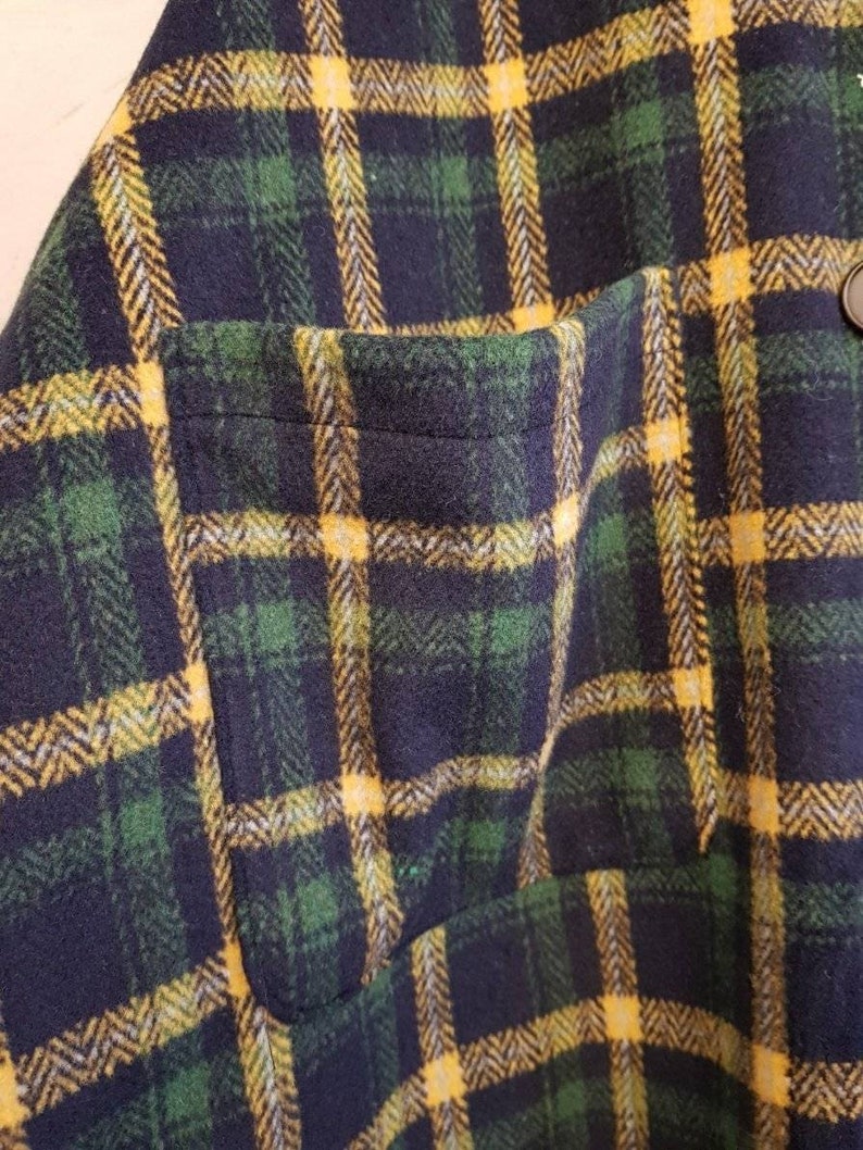 May include: A flannel shirt featuring a dark navy blue, forest green, and bright yellow plaid pattern. The garment includes a single patch pocket on the front and a dark button visible near the top edge of the pocket, showcasing a textured fabric.
