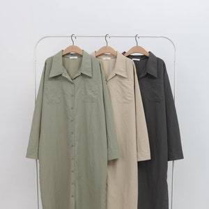 Op de afbeelding: Drie langarmige overhemden hangen aan een rek. De overhemden zijn in tinten groen, beige en grijs. Elk overhemd heeft een knoopsluiting, een kraag en twee zakken.