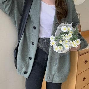 Op de afbeelding: Een lichtgroene cardigan met knoopsluiting en een zwarte leren tas over de schouder. De cardigan wordt gedragen over een wit T-shirt en een zwarte jeans.